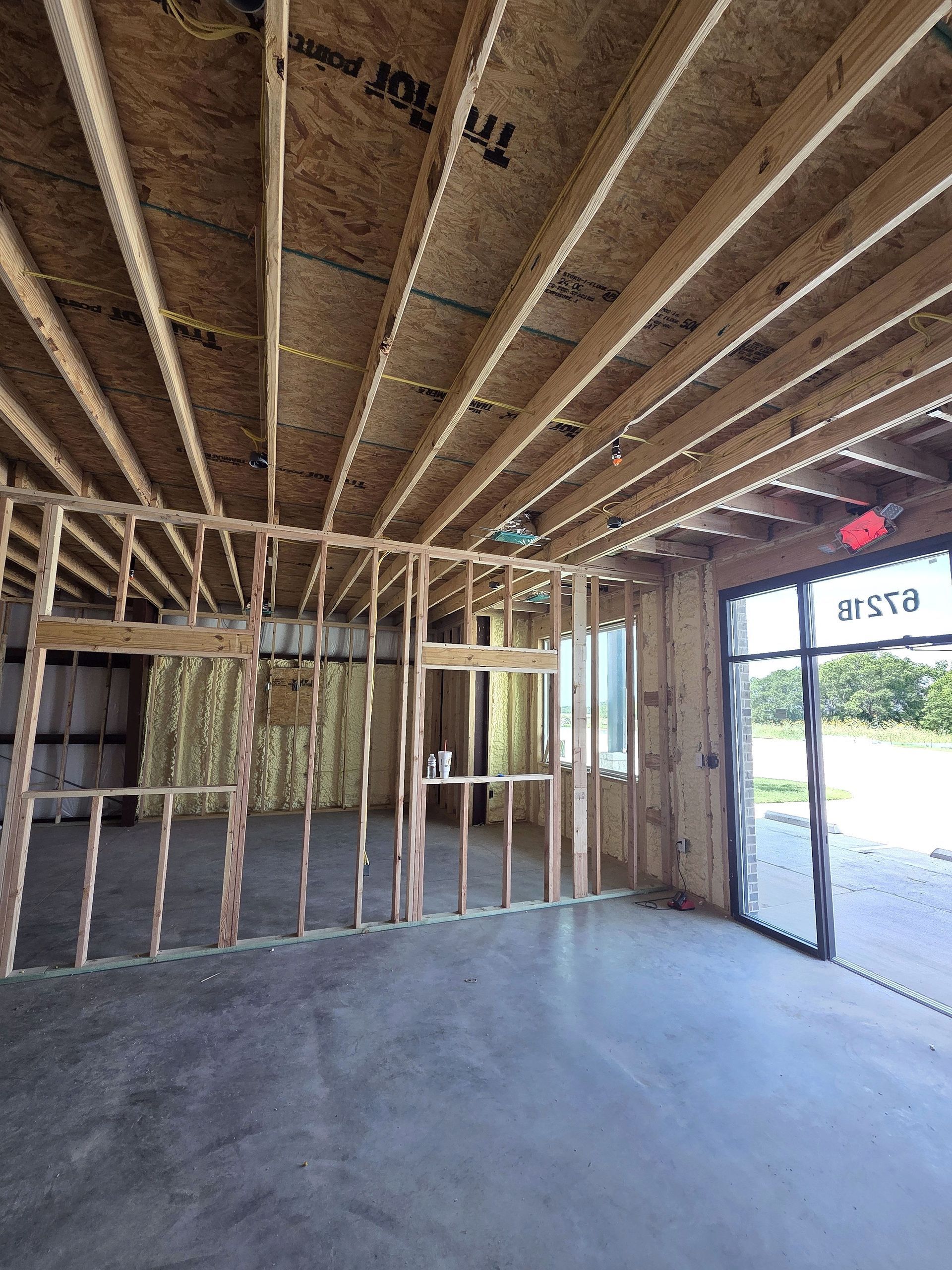 Interior view of a building under construction. Bare wooden framing for walls and ceiling, concrete floor, and an open doorway.