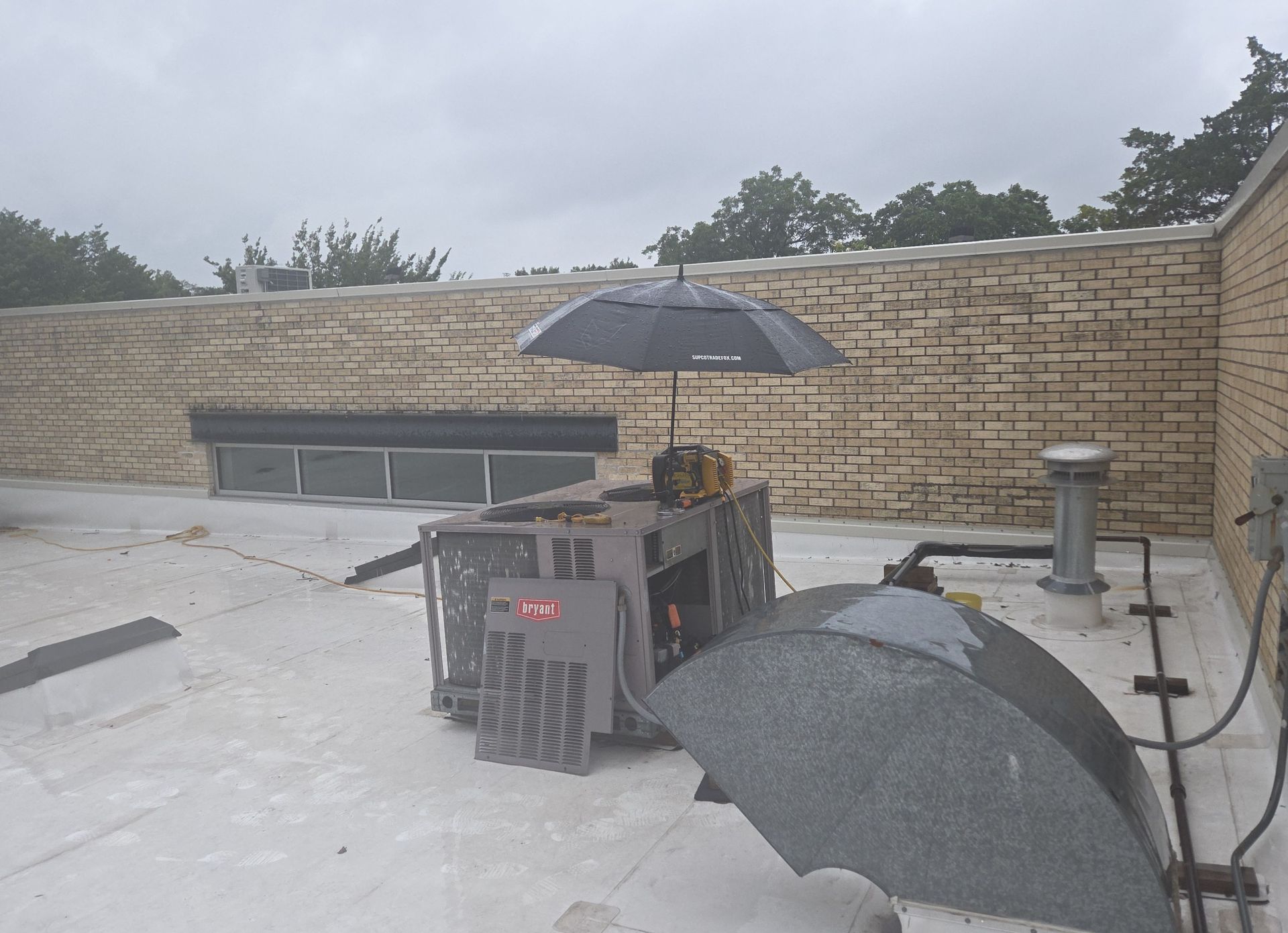 An open black umbrella is precariously perched atop an air conditioning unit on a flat rooftop, under a cloudy sky.