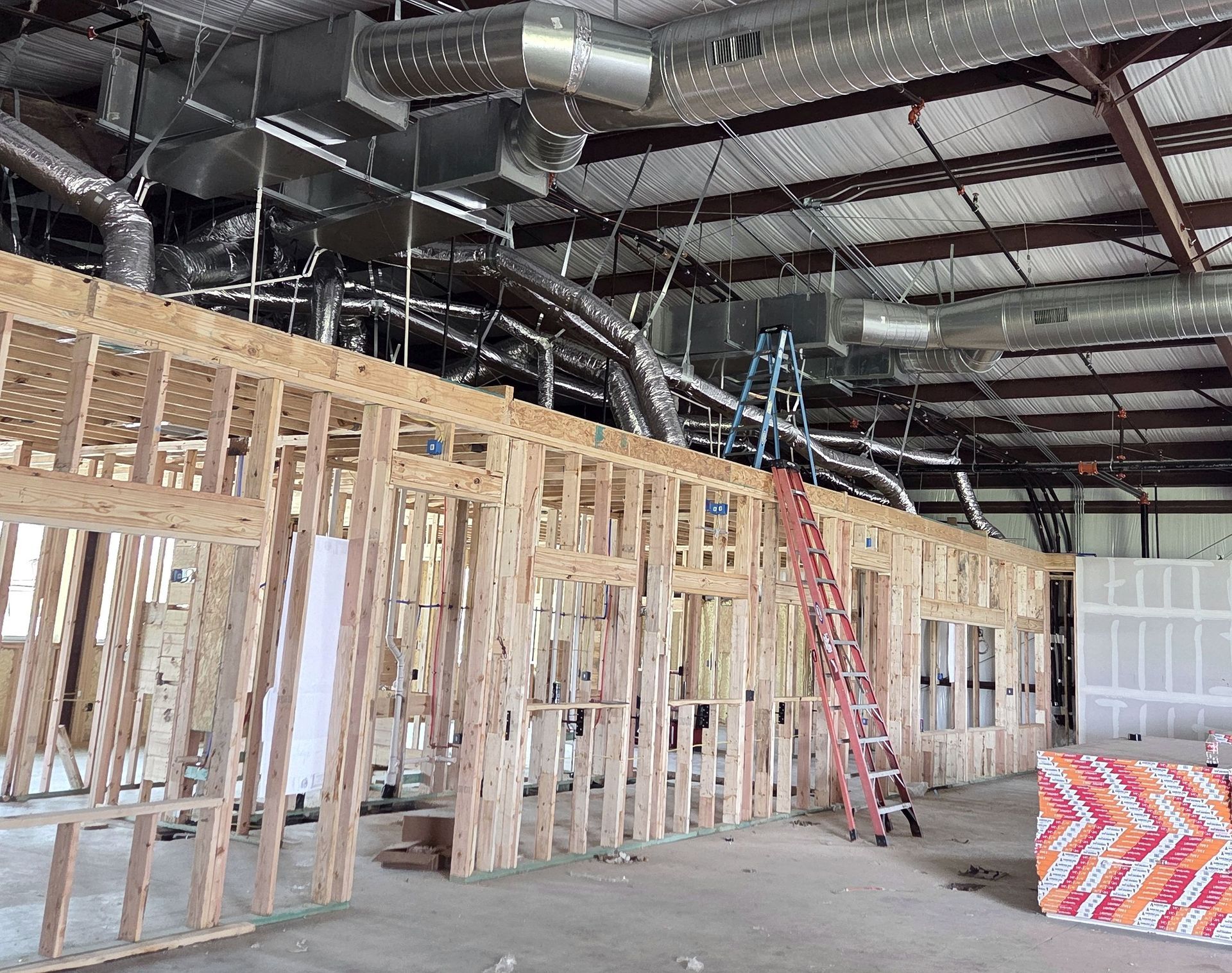 Interior view of a building under construction, with exposed wooden framing and ventilation ducts overhead. A ladder leans against the unfinished wall.