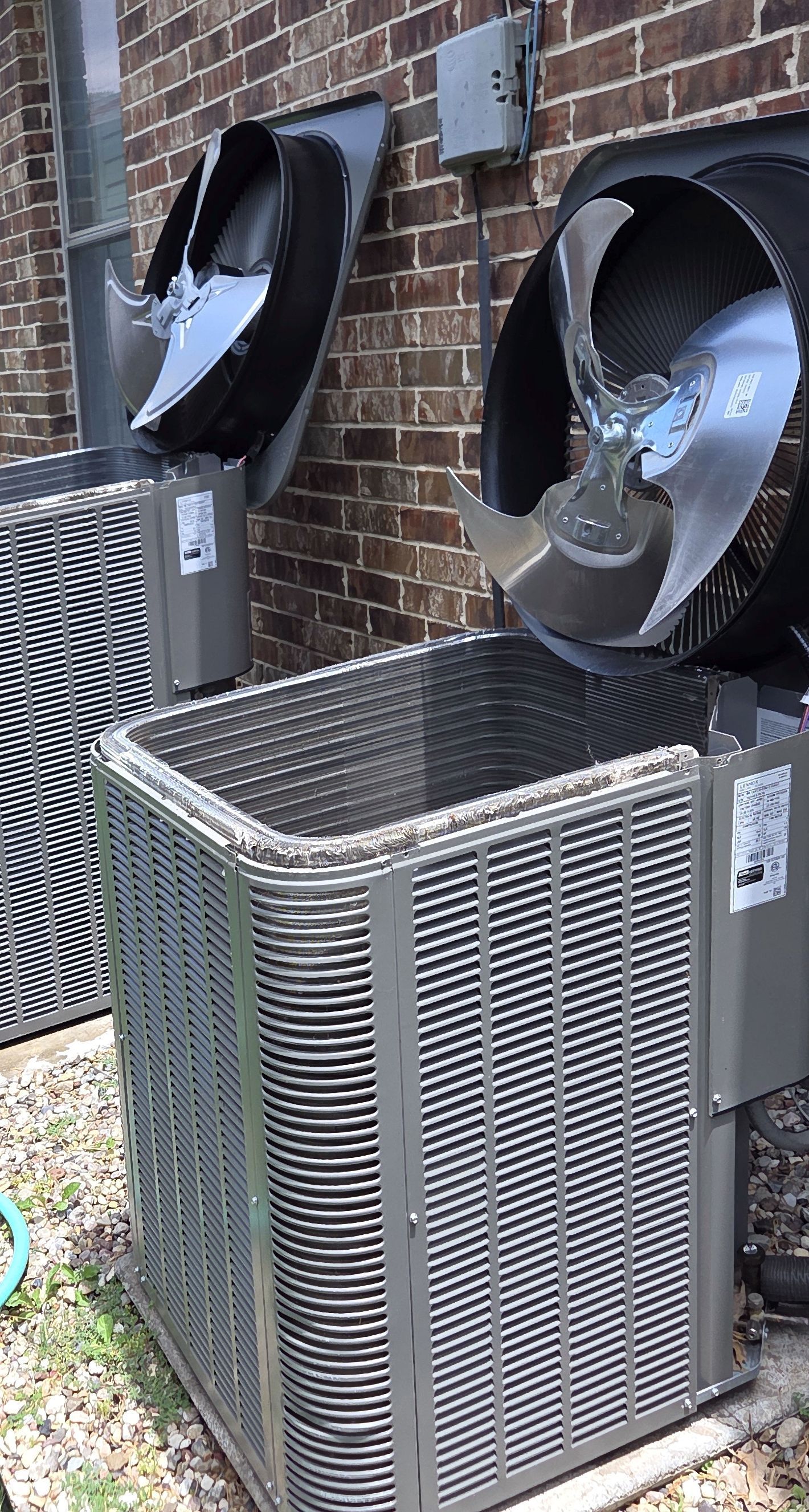 Two outdoor air conditioning units with fan blades visible, set against a brick wall.