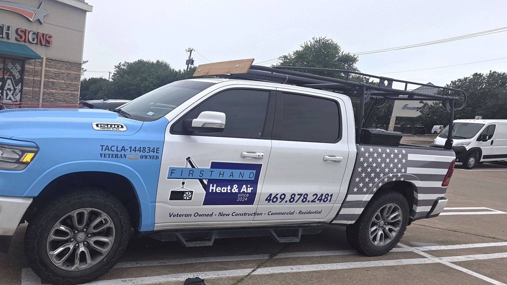 A light blue and white pickup truck with the American flag design parked outside a store.  The truck has a roof rack and carries the business name, 