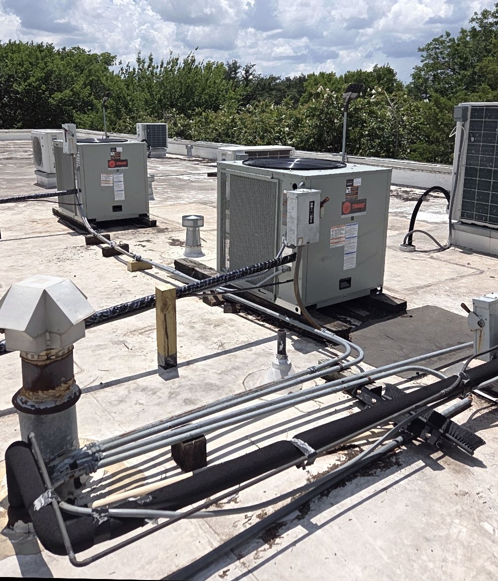 Rooftop with HVAC units, pipes, and vents. Gray machines sit on a white roof with trees in the background under a partly cloudy sky.