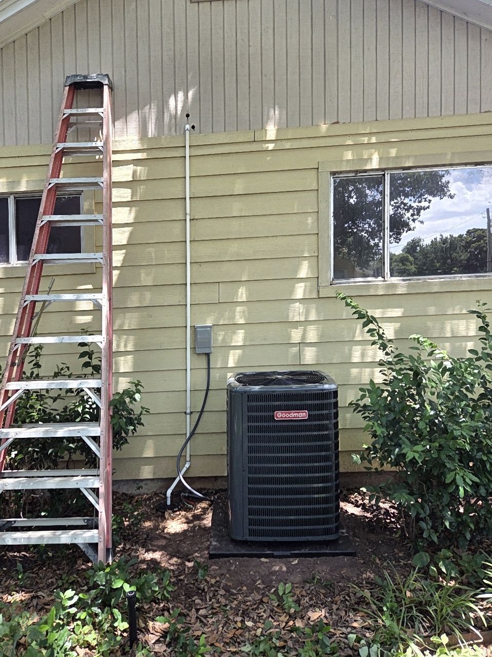 A tall red ladder leans against a yellow house, next to an air conditioning unit. A window and bushes are also in the view.