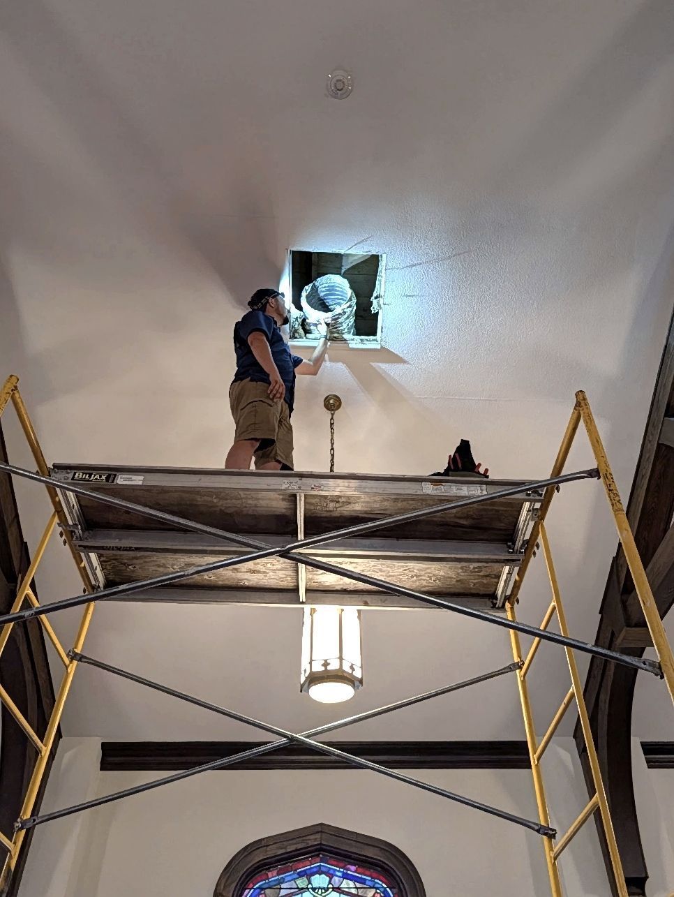 A person on a scaffold works on a ceiling fixture in a church. They reach into the opening with tools.
