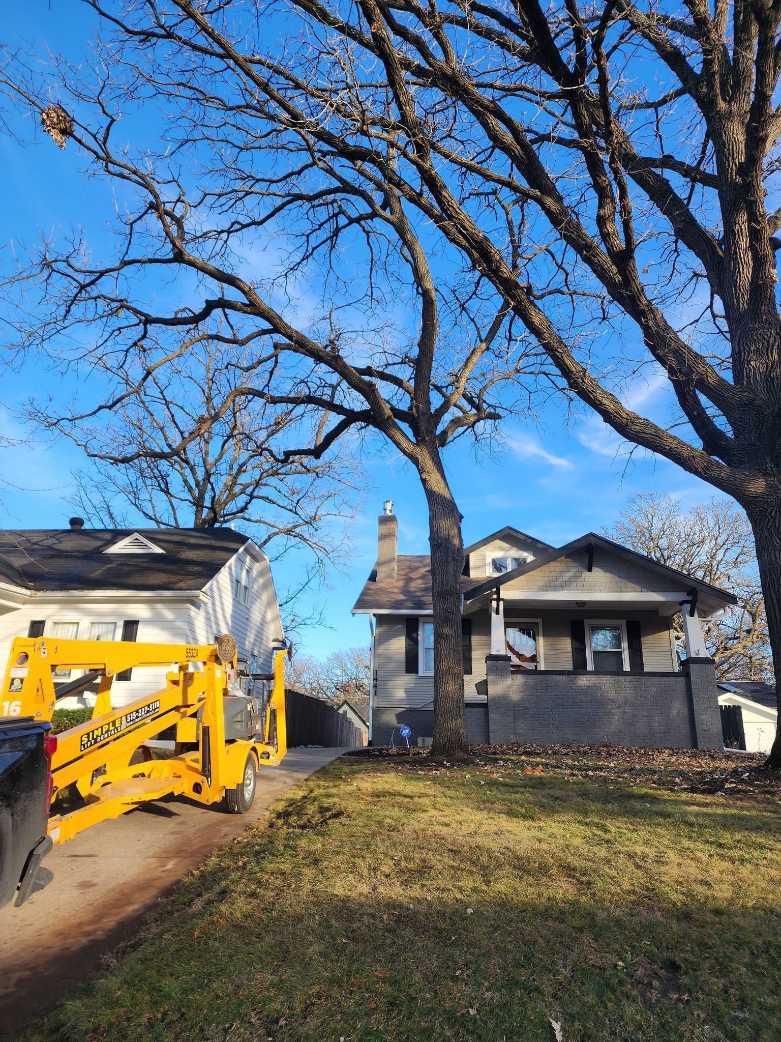 A yellow crane is cutting a tree in front of a house.