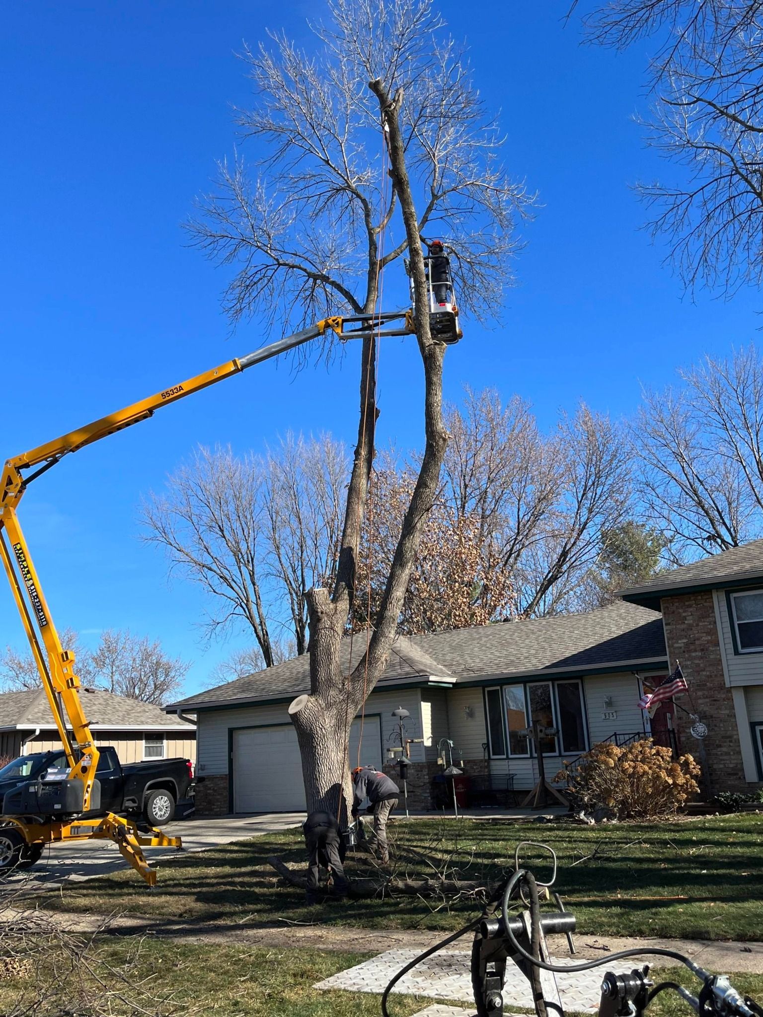 A tree being cut down by a crane in front of a house.