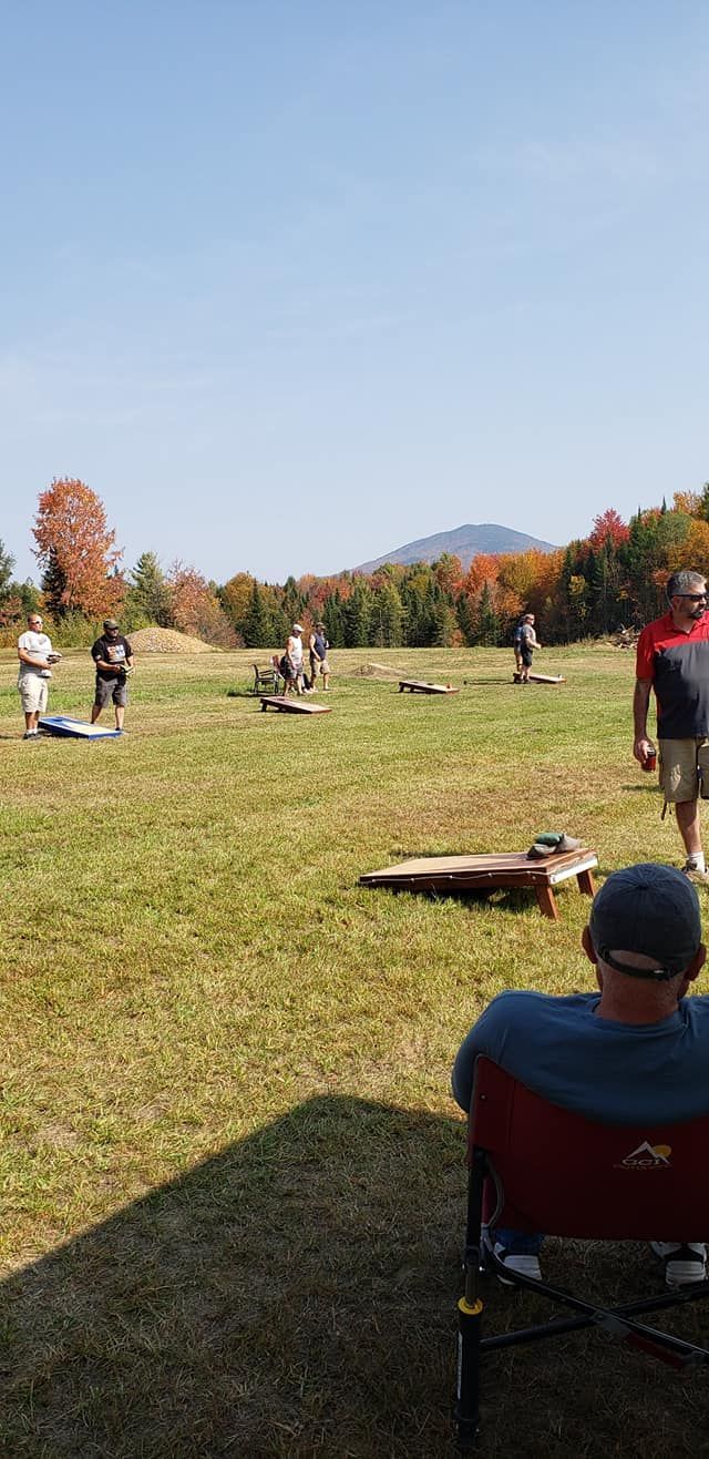 A man is sitting in a chair in a field watching people play cornhole.