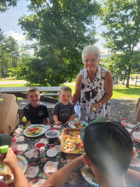 An elderly woman is standing next to three children at a picnic table.