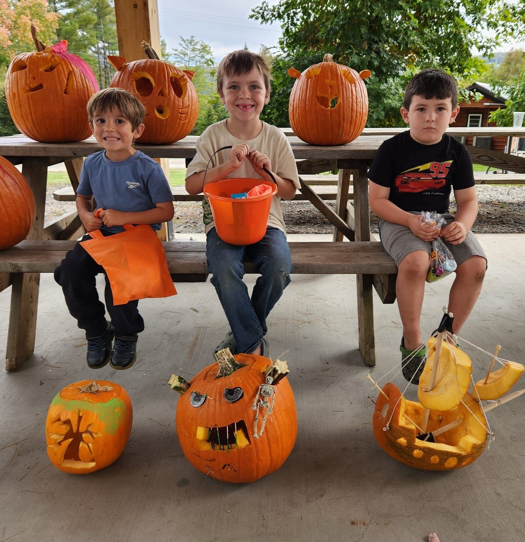 Three young boys are sitting on a picnic table surrounded by pumpkins
