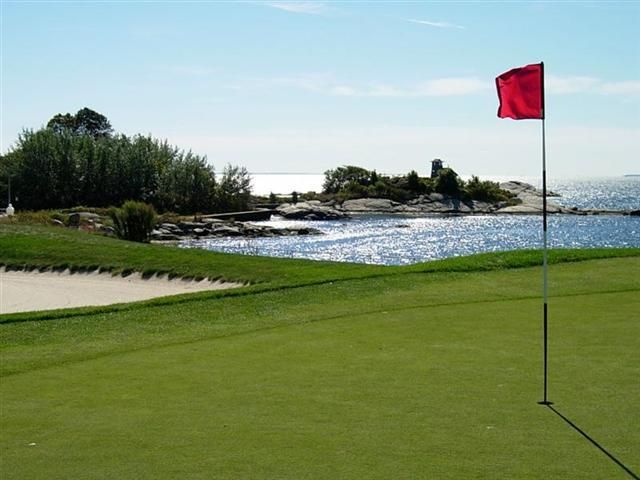 Golf green beside water with a red flag and sand bunker under a clear sky