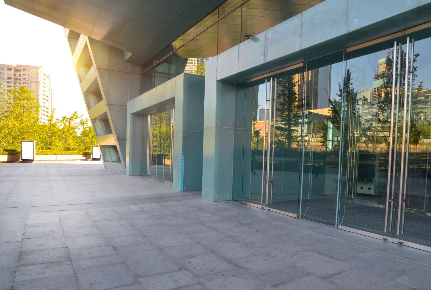 Modern building entrance with large glass windows and concrete pillars on a paved plaza, bathed in golden sunlight.