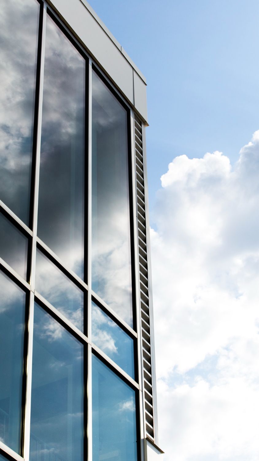 A modern office building corner with reflective glass windows mirroring the blue sky and clouds.