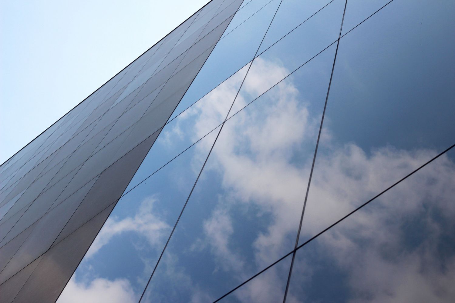 Low-angle view of a modern building facade with reflective glass windows mirroring a bright blue sky and white clouds.