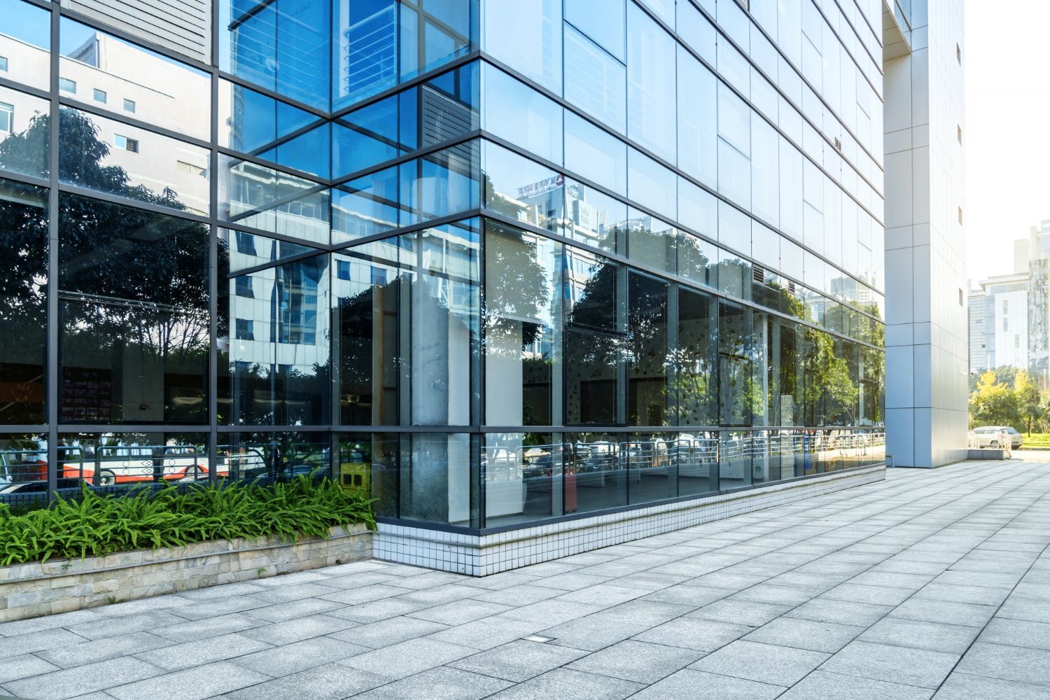 A modern glass-paneled office building with a paved outdoor plaza and greenery at its base.