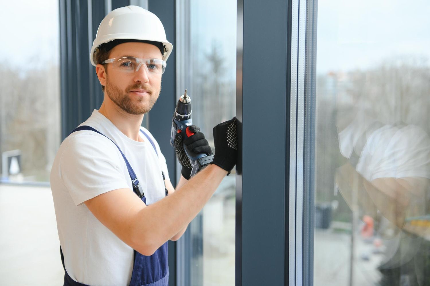 A worker in a white hard hat and safety glasses holding a power drill by a large glass window frame.