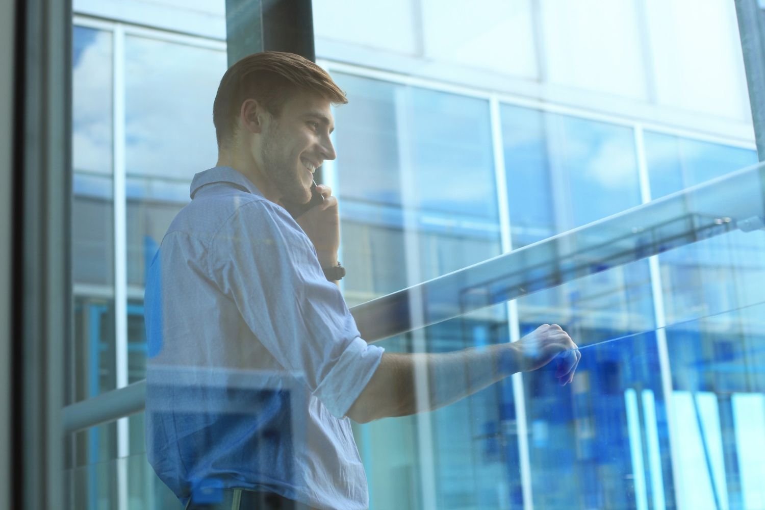 A smiling person in a light shirt speaks on a phone while standing by a glass office building wall.