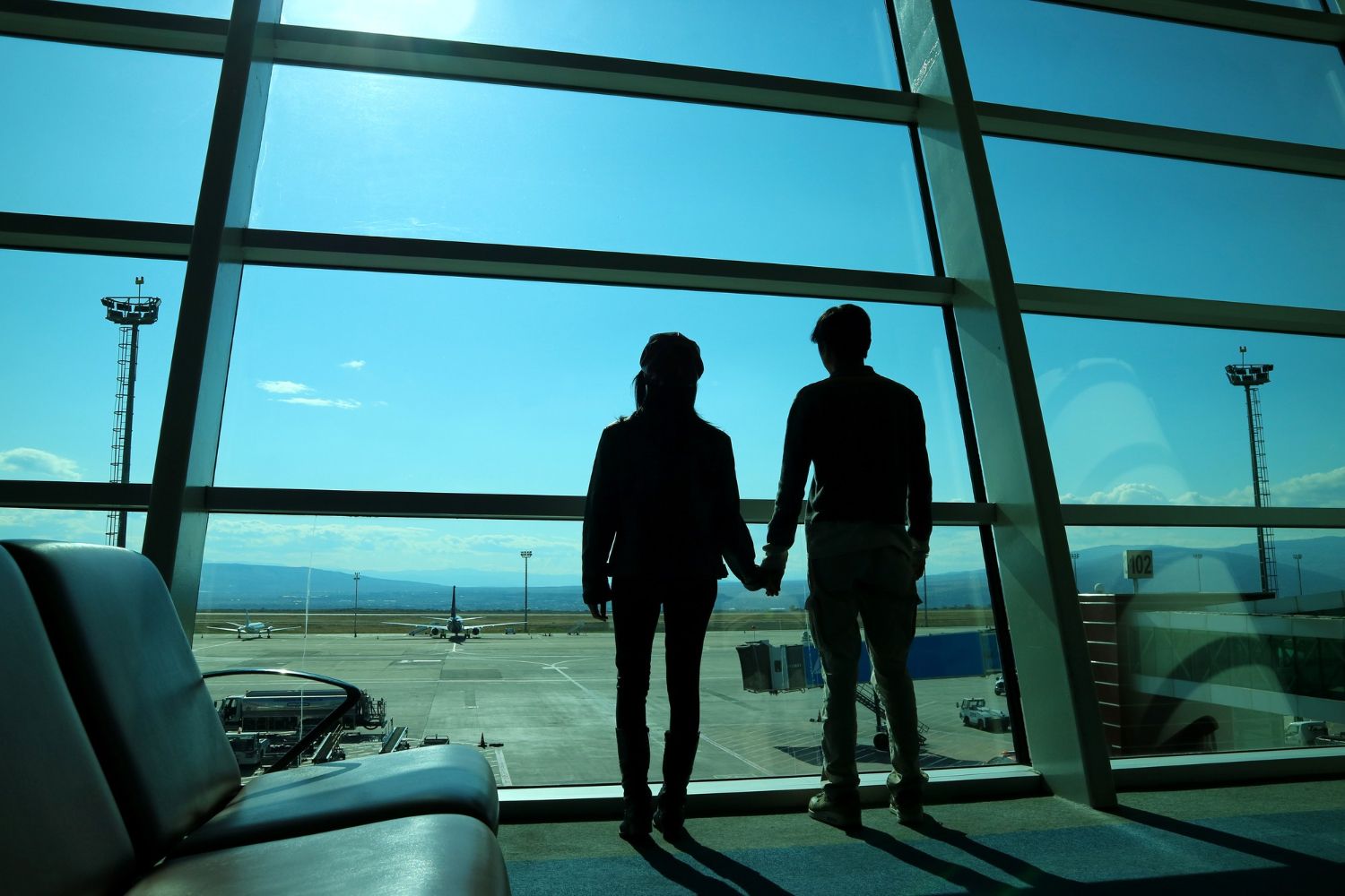 Two people hold hands while looking out a large airport window at an airplane on the tarmac.