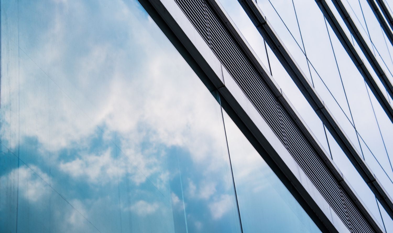 A modern glass building facade features blue, cloud-filled reflections next to angled steel supports and metal mesh.