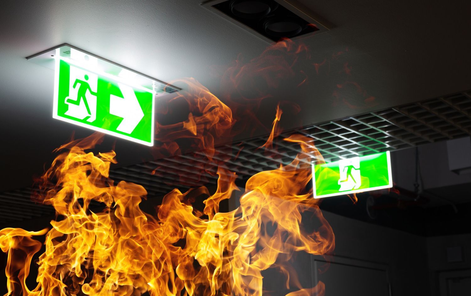 Two illuminated green exit signs mounted on a ceiling above a large, bright orange fire.