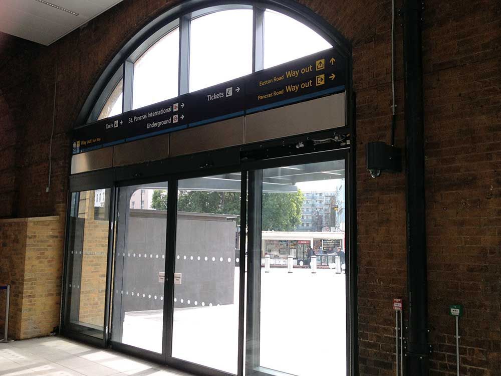Sliding glass doors set in a brick wall at a transit station, with overhead signage and an arched window above.