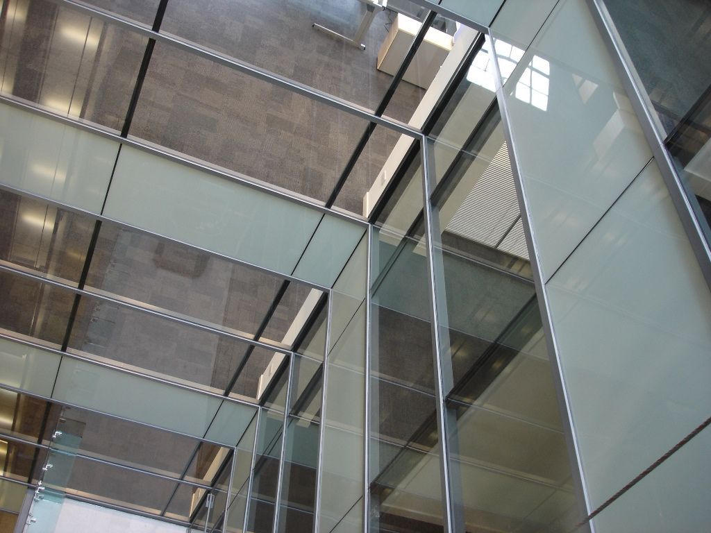 A low-angle view of a modern glass building interior, highlighting geometric glass panels, metal framing, and a ceiling.