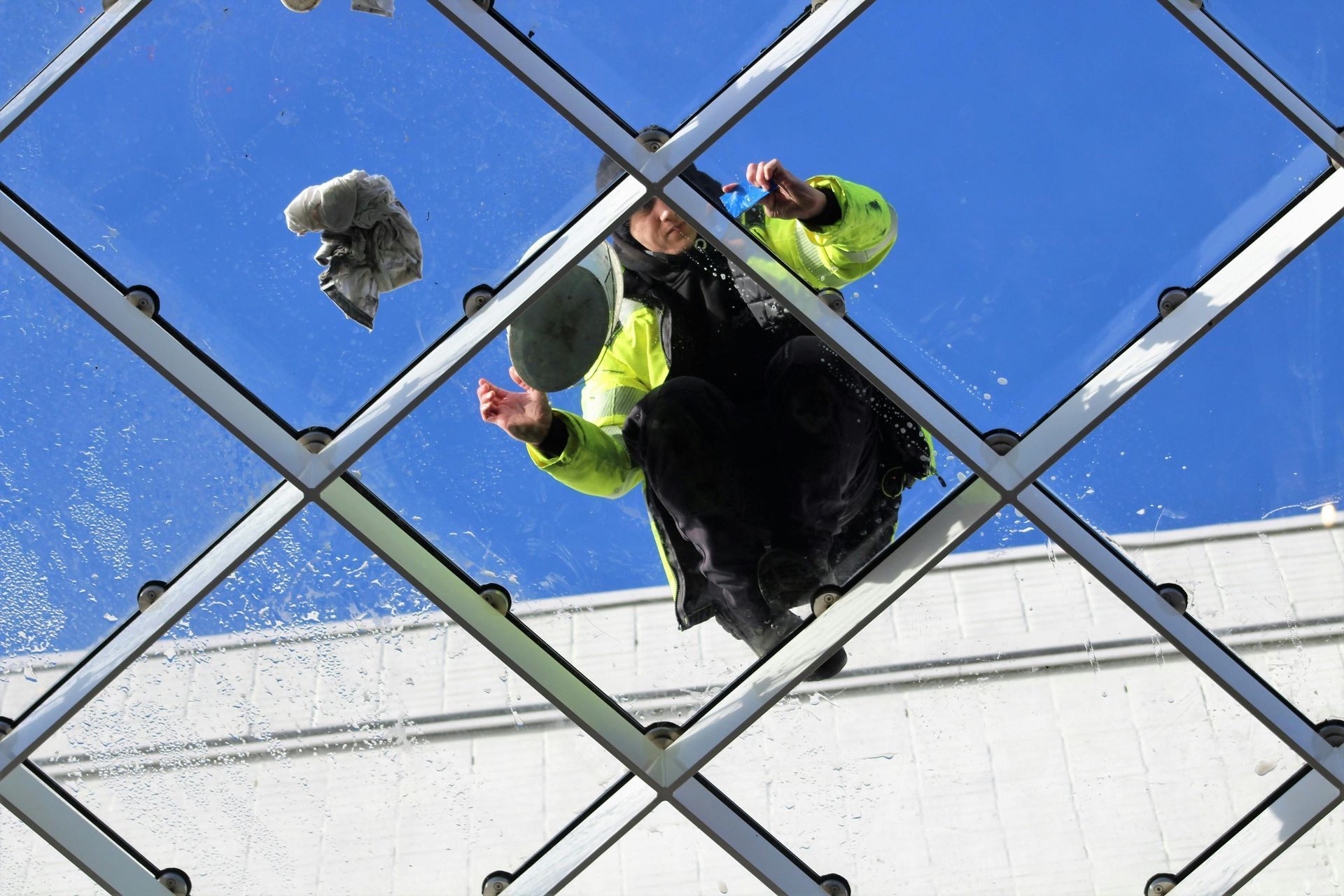 A worker in a bright yellow high-visibility jacket cleans a glass ceiling from above, seen through the structural frame.