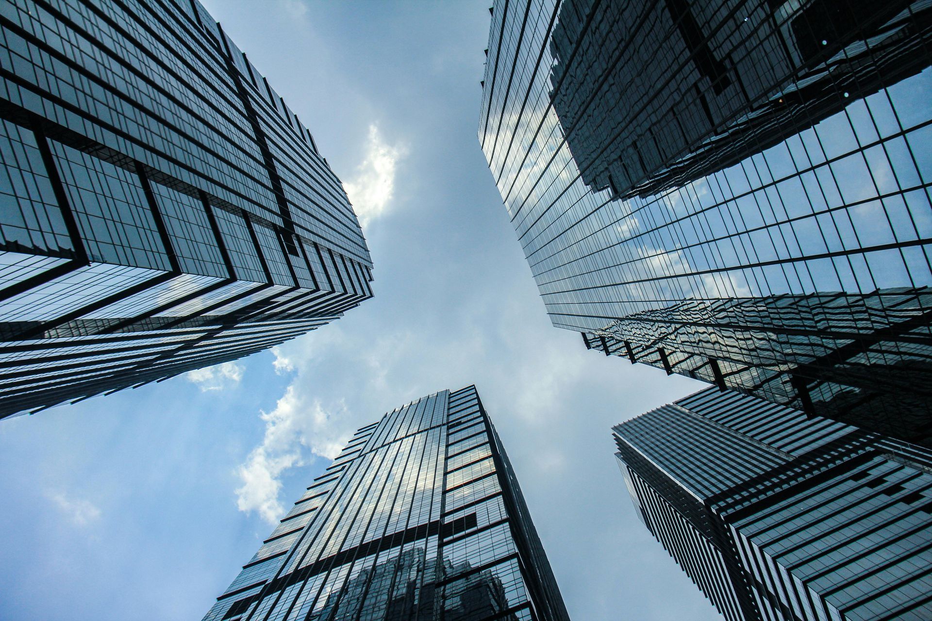 Low-angle view of several modern glass skyscrapers reaching toward a cloudy blue sky.