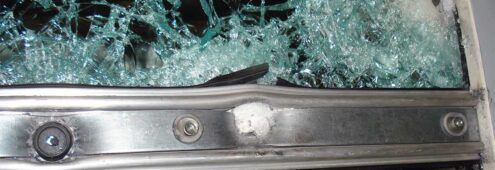 Shattered glass pane held within a metal frame featuring visible scuffs, a central white deposit, and mounting screws.