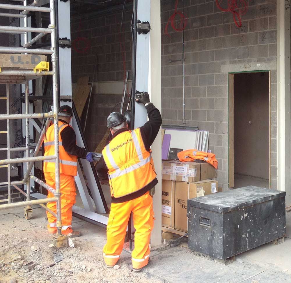 Two workers in high-visibility orange safety gear install a panel on a building construction site.