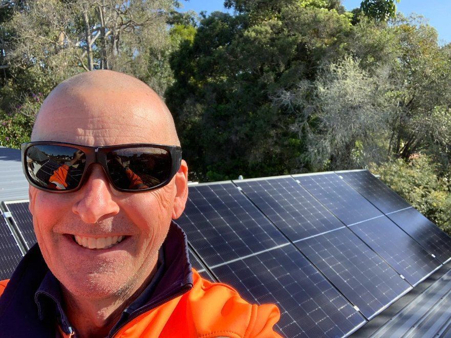 A Man Wearing Sunglasses is Smiling in Front of a Row of Solar Panels — Dylko Electrical in Wauchope, NSW