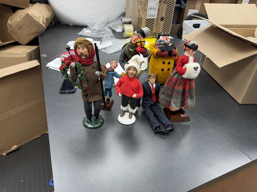 A group of six small holiday figurines stand on a gray surface surrounded by cardboard boxes in a workshop setting.