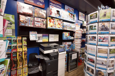 A retail store interior features shelves stocked with toys and games, a photocopier, and a rotating rack of greeting cards.