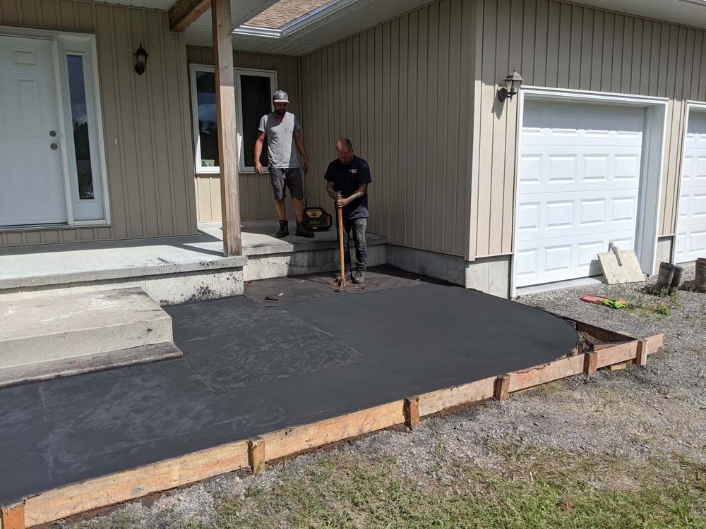 Two men are working on a driveway in front of a house.