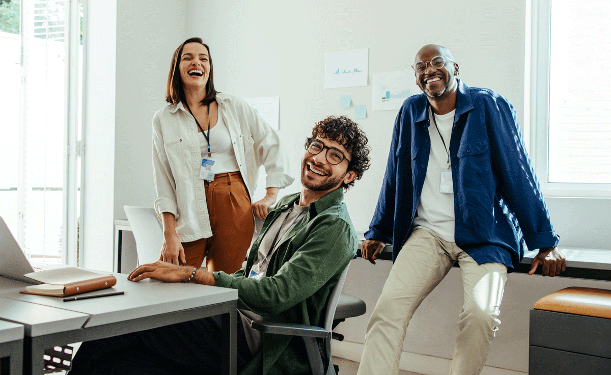 Three smiling people pose in an office. One sits at a desk, two lean against a wall.