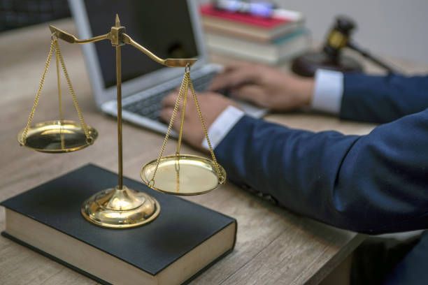Attorney working on laptop with justice scale on thick law book and gavel on desk in the background.