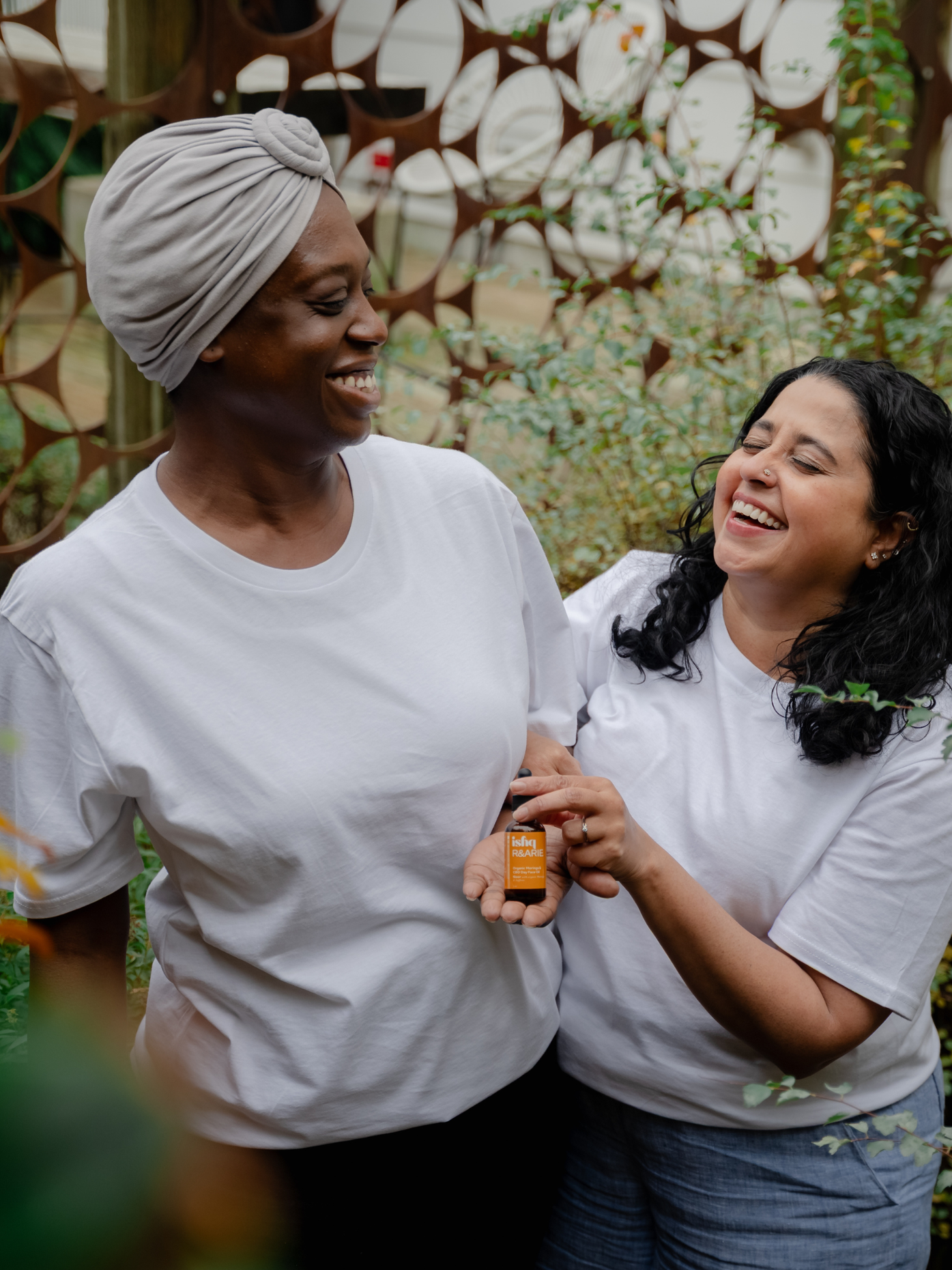 Two women-one black & the other brown are laughing and holding a one ounce bottle of Ishq Organic CBD-Moringa Day Face Oil. The background is plants, and they are standing in the middle. This oil is magic for the skin. it smoothes dry skin, hydrates deeply, builds skin's barrier, brings a glow to the face. Named Noor, which means Light in Urdu & Trinidadian, this is a collaboration between Ishq and R&ARIE. 