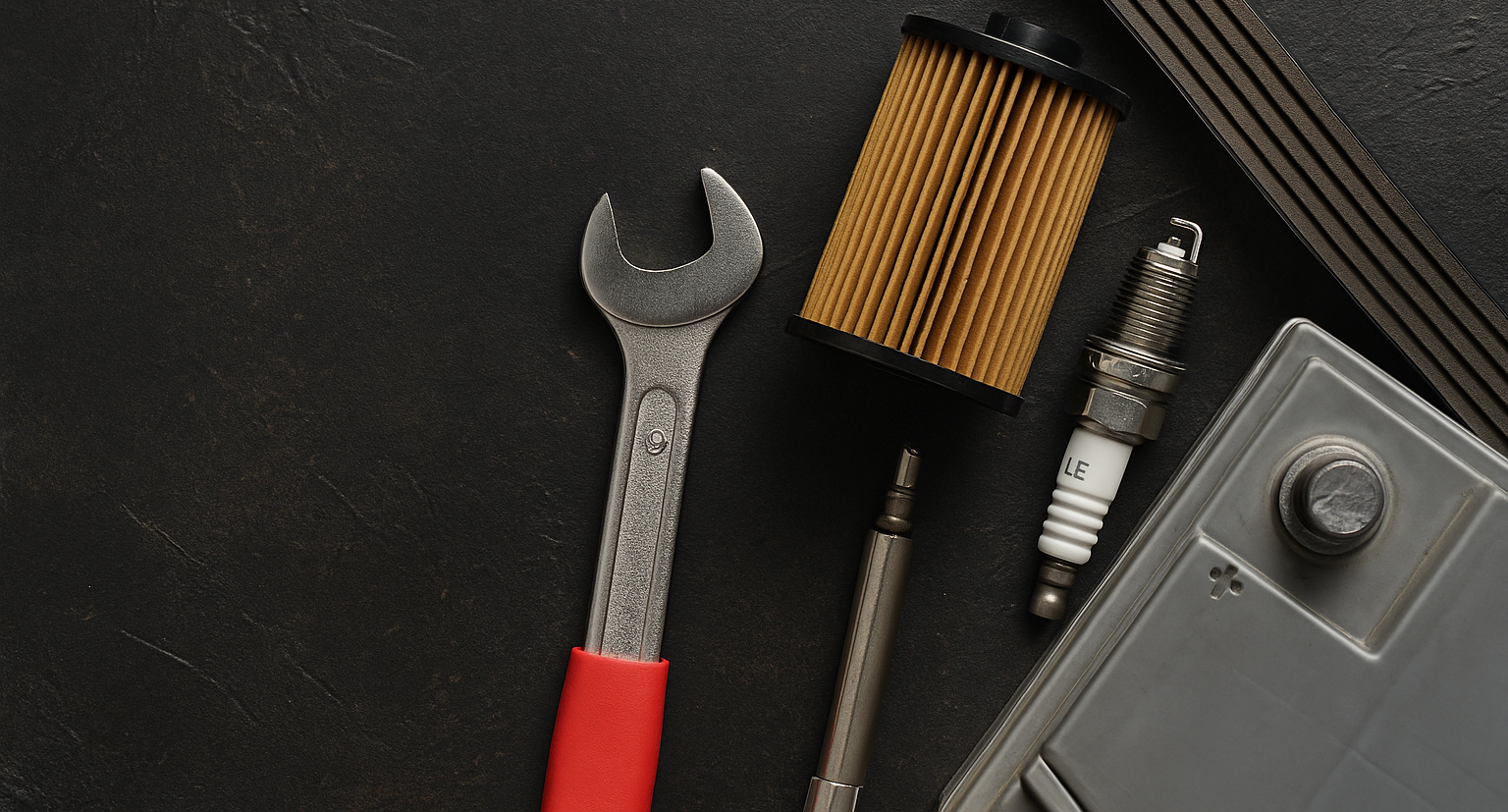 Close-up of essential car maintenance tools laid out on a dark textured surface, including a wrench with a red handle, an oil filter, a spark plug, a multi-ribbed serpentine belt, and a car battery corner—ideal for illustrating scheduled automotive service concepts.