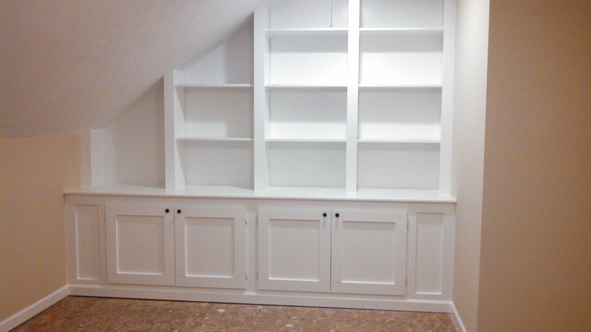 A room with a slanted ceiling and white shelves and cabinets.