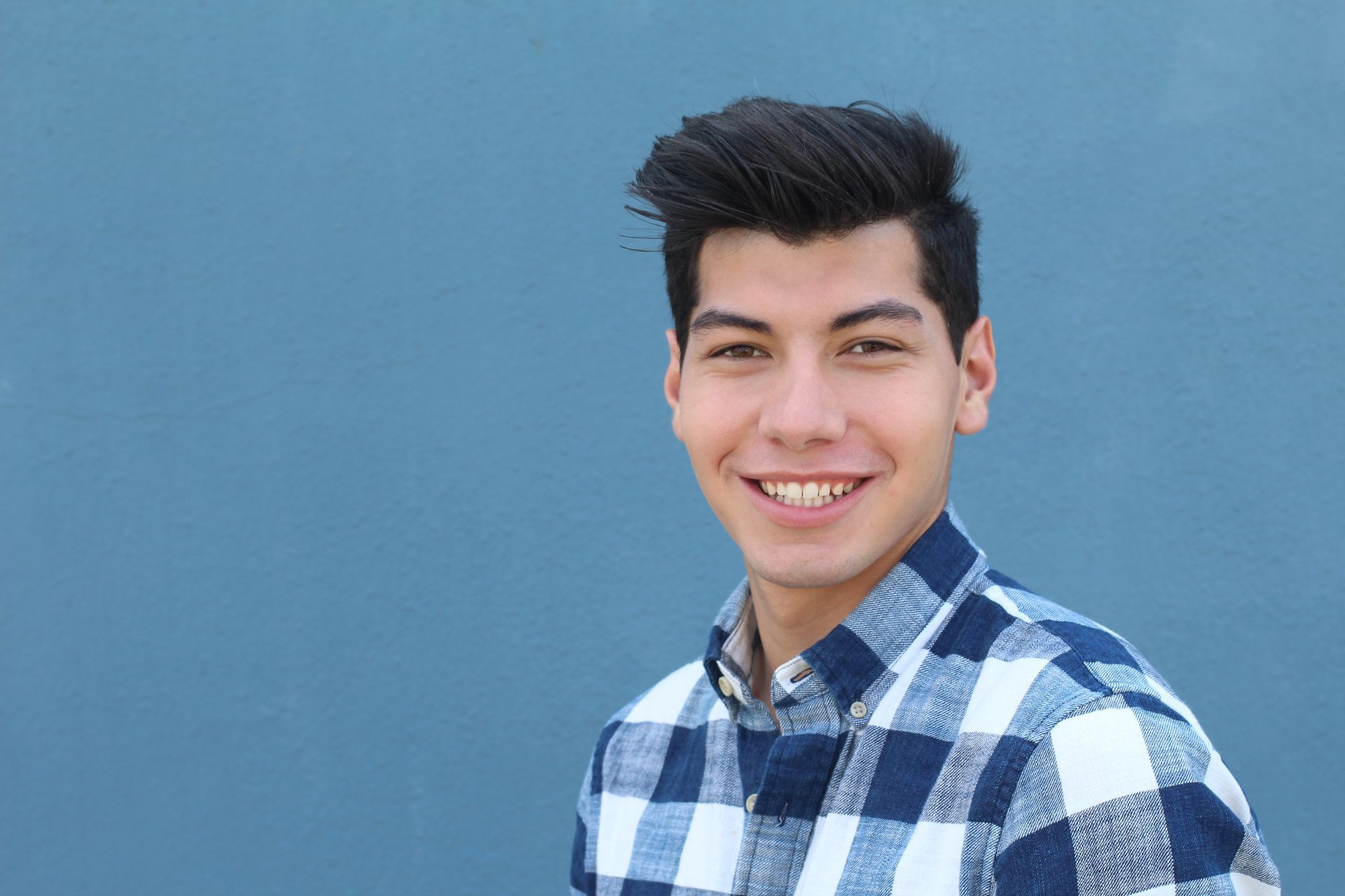 A young man in a plaid shirt is smiling in front of a blue wall.