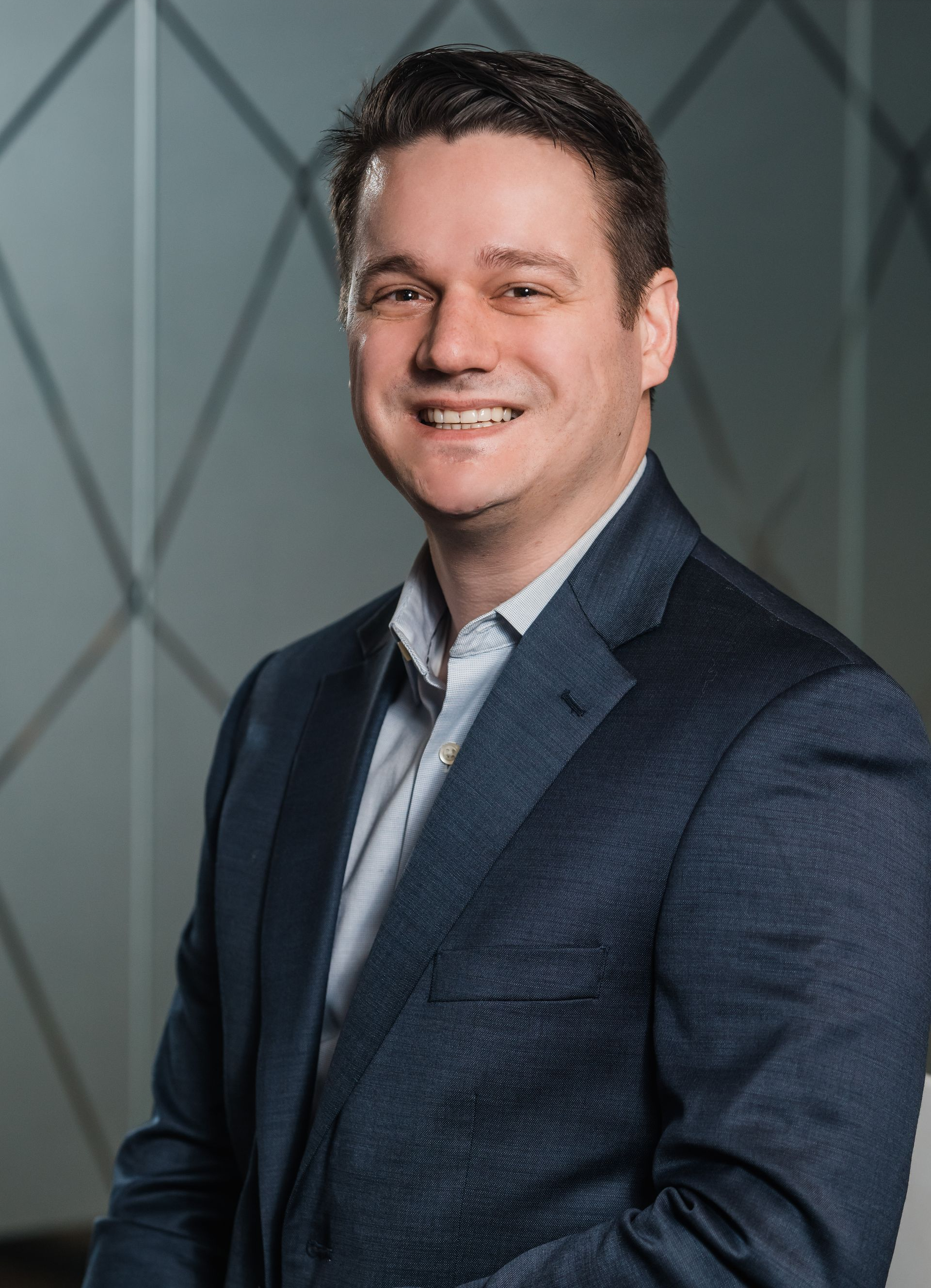 Man in a blue shirt and navy blazer smiling at the camera against a white background.