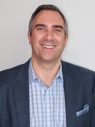 Man smiling, wearing a blue blazer over a light blue plaid shirt, and pocket square.