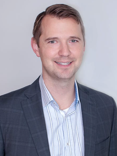 Man in blue striped shirt and gray blazer smiles at the camera, against a light gray background.