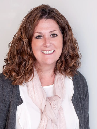 Woman with curly brown hair, smiling, wearing a white shirt, pink scarf, and gray cardigan in front of a light gray wall.