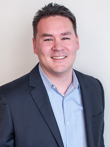 Man in a blue shirt and navy blazer smiling at the camera against a white background.