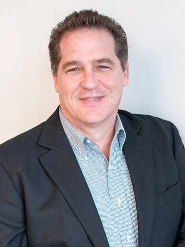 Man in blazer smiles at the camera, light blue collared shirt.