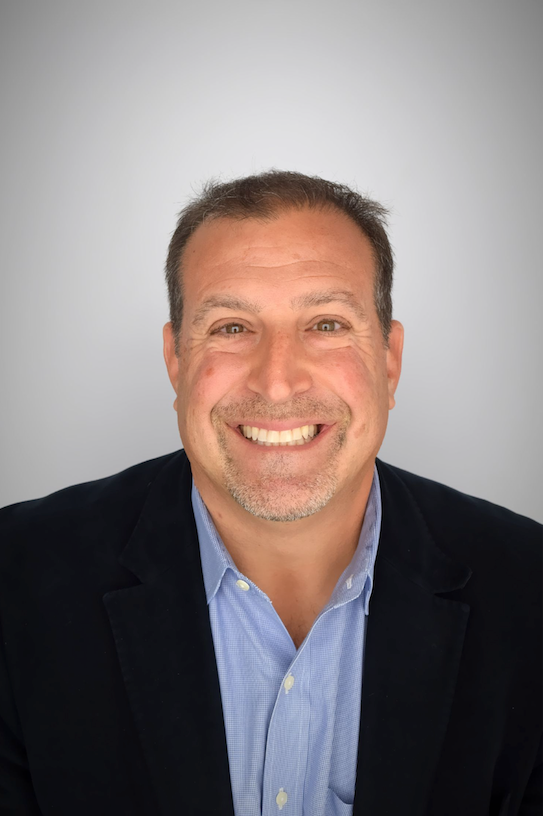 Man smiling, wearing a blue shirt and navy blazer against a gray background.