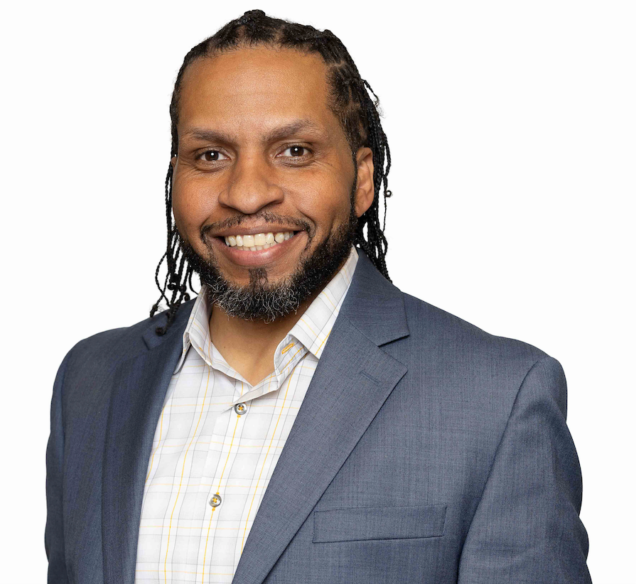 Man with dreadlocks smiling, wearing a blue blazer and light patterned shirt.