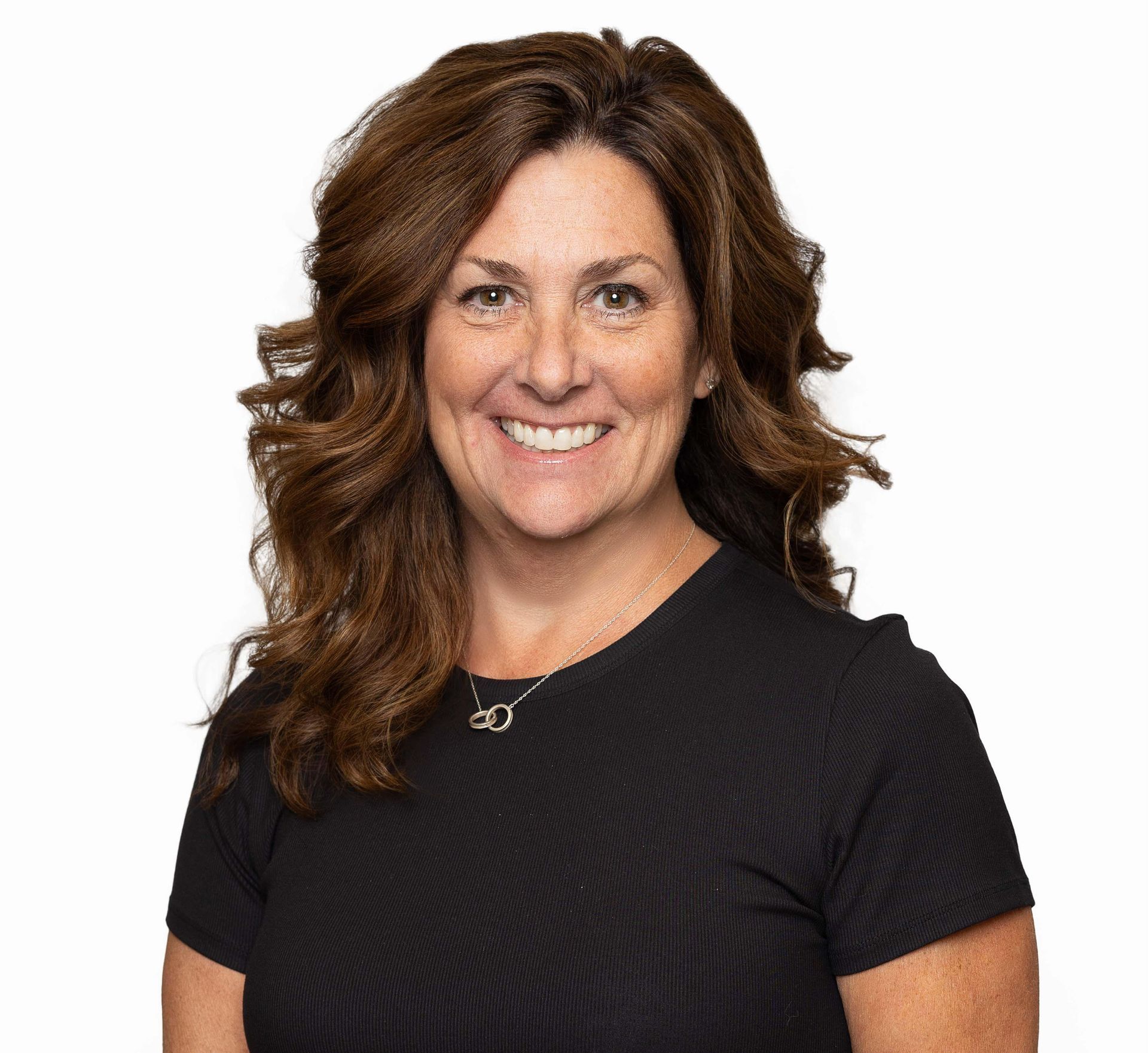 Woman with curly brown hair, smiling, wearing a white shirt, pink scarf, and gray cardigan in front of a light gray wall.
