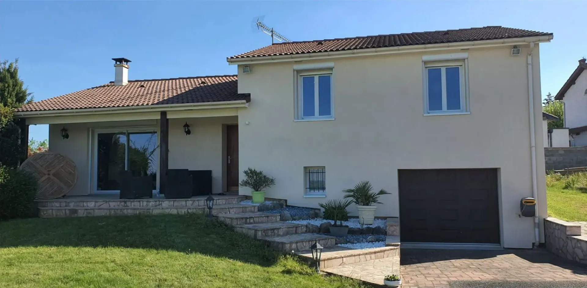 Une maison claire de deux étages avec un toit de tuiles brunes, un garage intégré et une terrasse surélevée, par une journée ensoleillée.