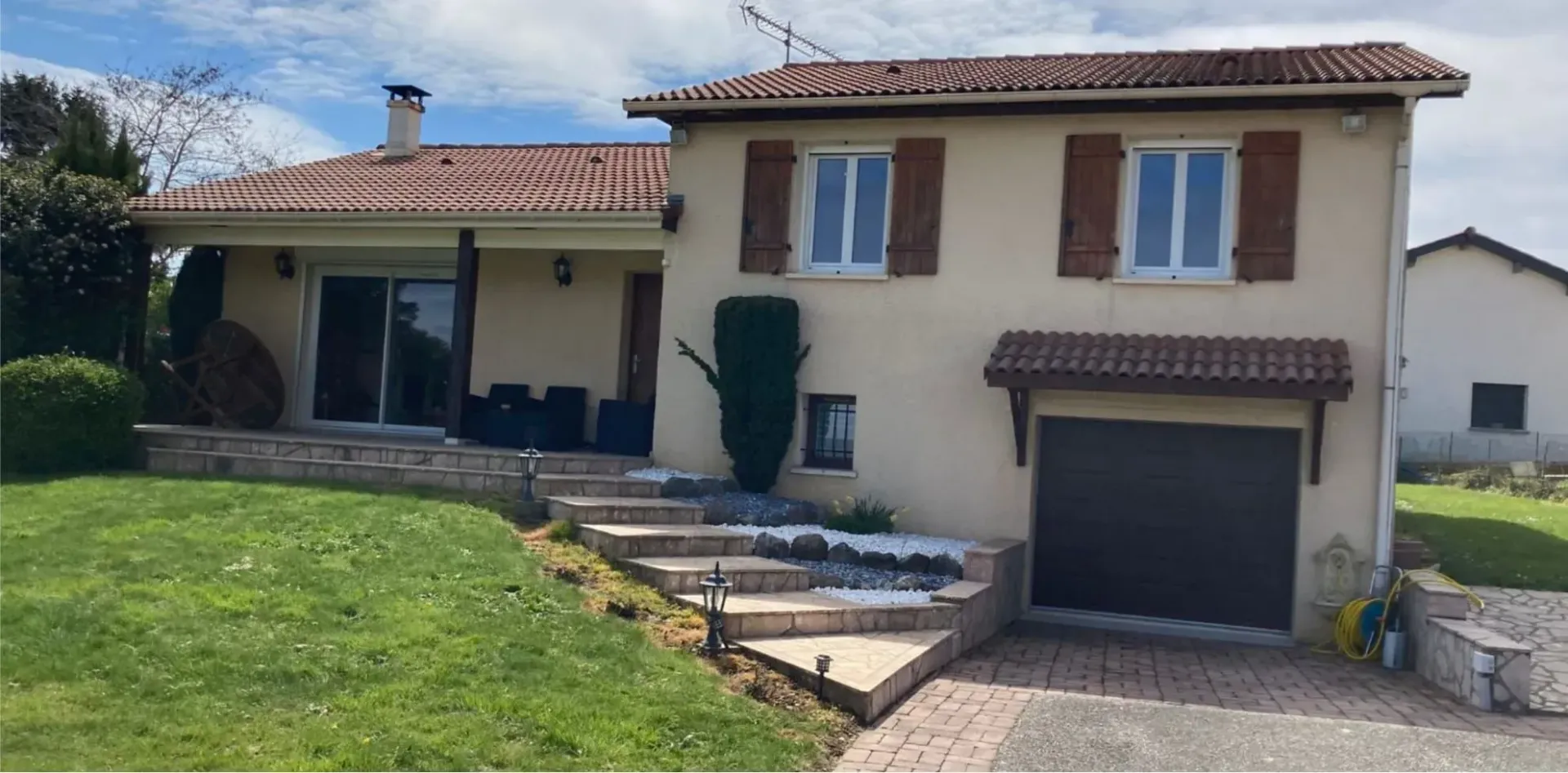 Une maison à deux étages en stuc avec un toit de tuiles, des volets en bois, un garage et des marches en pierre menant à une terrasse surélevée.